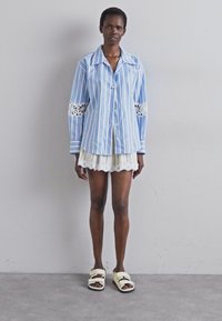 Stripet blå knapp-up skjorte med blonde detaljer på ermene, kombinert med et lyst plissete skjørt og beige sandalsko. Jevn stofftekstur.