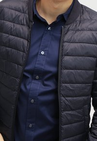 Man med en marinblå knäppt skjorta och en svart vadderad jacka med dragkedja, sedd från halsen till midjan.