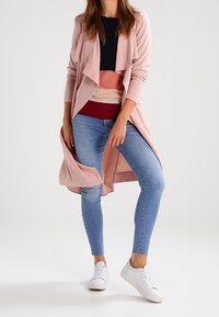 Długi, jasnoróżowy kardigan z drapowanym przodem, granatowa bluzka i warstwowe jasne kolory. Dopełnieniem stroju są dopasowane niebieskie dżinsy i białe trampki.