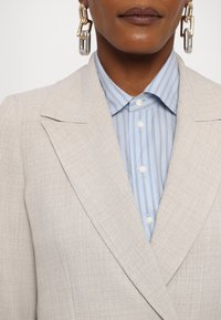 Lys beige blazer med teksturert stoff og fremtredende slag, over en lys blå skjorte med knapper og tynne hvite striper. Store, metalliske kjedeøredobber.