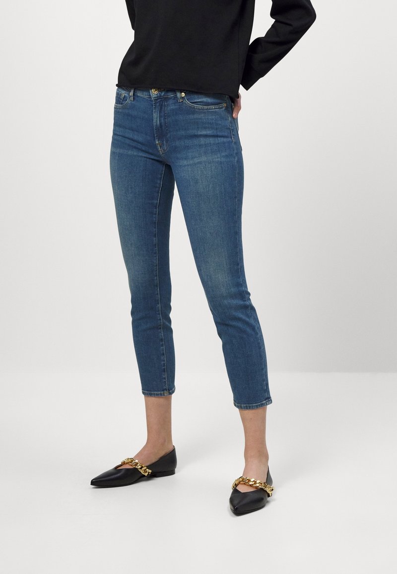 Skinny jeans in medium wash denim, cropped above the ankle. Paired with black flats featuring a gold chain accent.
