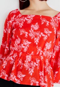 Czerwona bluzka peplum z kwadratowym dekoltem i długimi rękawami, z różowymi wzorami kwiatowymi na teksturowanej tkaninie.