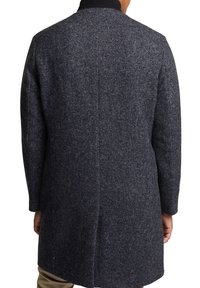 Uomo che indossa un cappotto di lana a spina di pesce grigio scuro visto da dietro, abbinato a pantaloni beige e a un dolcevita nero.