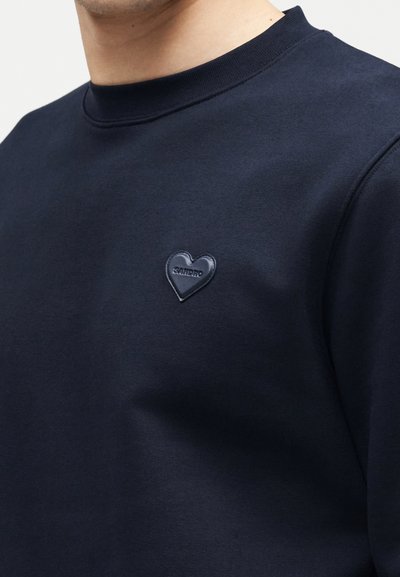 Άνδρας που φοράει μπλε σκούρο φούτερ με μικρό μαύρο καρδιόσχημο κεντητό μπαλώμα στο στήθος με το όνομα της μάρκας «Santero».