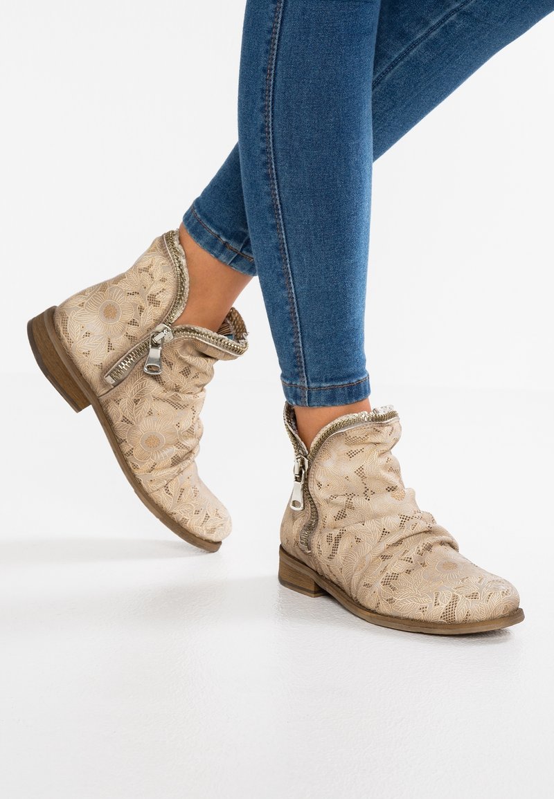 Bottes beige jusqu'à la cheville avec motif floral embossé, design décontracté, fermetures éclair latérales et petits talons en bois, associées à un jean bleu foncé.