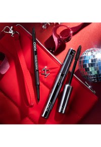 Tre KIKO skjønnhetsprodukter: en svart filtippen-eyeliner, et sølvfarget mascara-rør, og en svart mascara-børste, arrangert på en rød teksturert overflate.