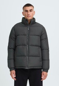 Mörkgrå puffjacka med hög krage, plisserad design, framzipper och elastiska ärmar. Har en slät, matt yta.