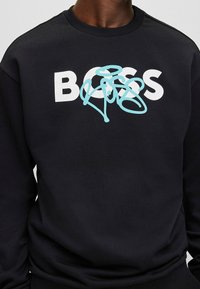 Sudadera negra con "BOSS" en blanco, con un diseño abstracto azul superpuesto. Hecha de tela suave con puños y cuello de canalé.