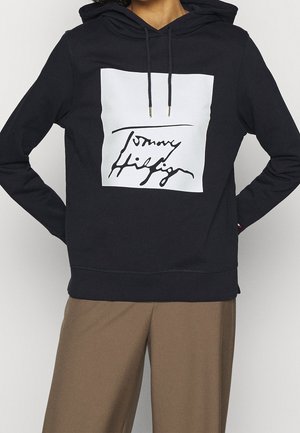 Persoon draagt een zwarte hoodie met een witte vierkant en zwarte cursieve tekst aan de voorkant, gecombineerd met een bruine broek, handen in de zij.