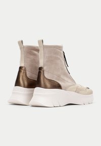 Botas de tobillo beige con parte superior de ante texturizado, acentos metálicos en bronce, panel lateral elástico y suela gruesa blanca. Presenta un cierre con cremallera.