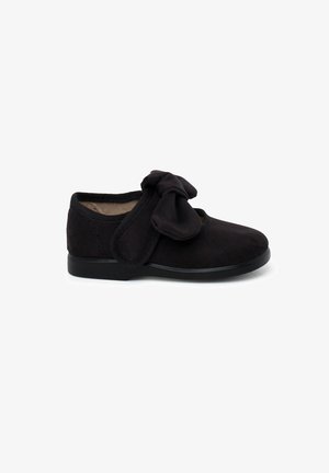 Zapato de ante negro con punta redonda, bordes elásticos y un lazo de tela en la parte frontal. Textura suave y suela de goma plana.