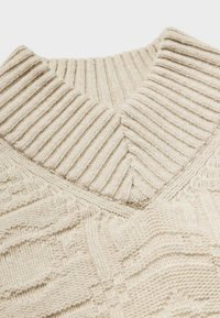 Pull en tricot beige avec un col côtelé et des motifs texturés, arborant un design en chevrons distinctif et une laine douce et épaisse.
