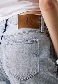 Vaqueros de mezclilla azul claro con una etiqueta de cuero marrón en la parte trasera de la cintura, grabada con "Calvin Klein Jeans" y un logotipo estilizado.
