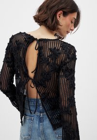 Zwarte doorschijnende top met bloemenborduursels, open rugontwerp, lange mouwen en striksluitingen. Geribbeld en textuurstof, gecombineerd met denim.