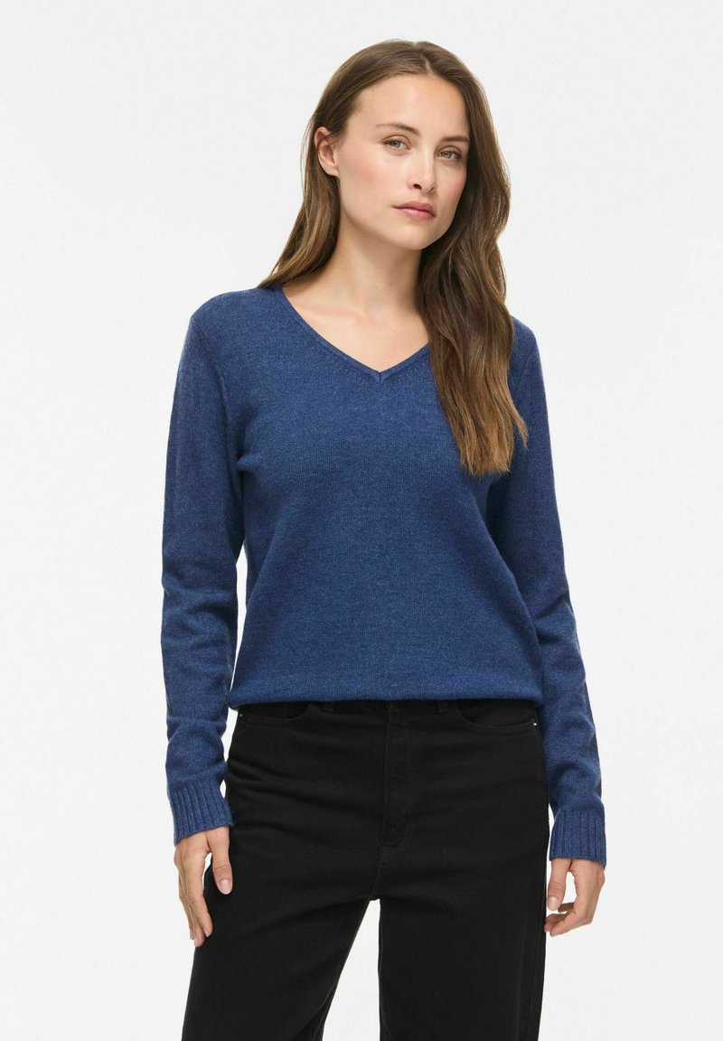 Femme aux longs cheveux bruns portant un pull bleu à col en V et un pantalon noir, debout sur un fond blanc.