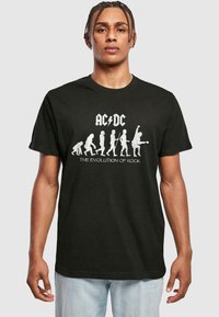 Svart t-skjorte med teksten "AC/DC" og "The Evolution of Rock" i hvitt, med grafiske silhuetter som viser menneskelig evolusjon til rockemusikere.