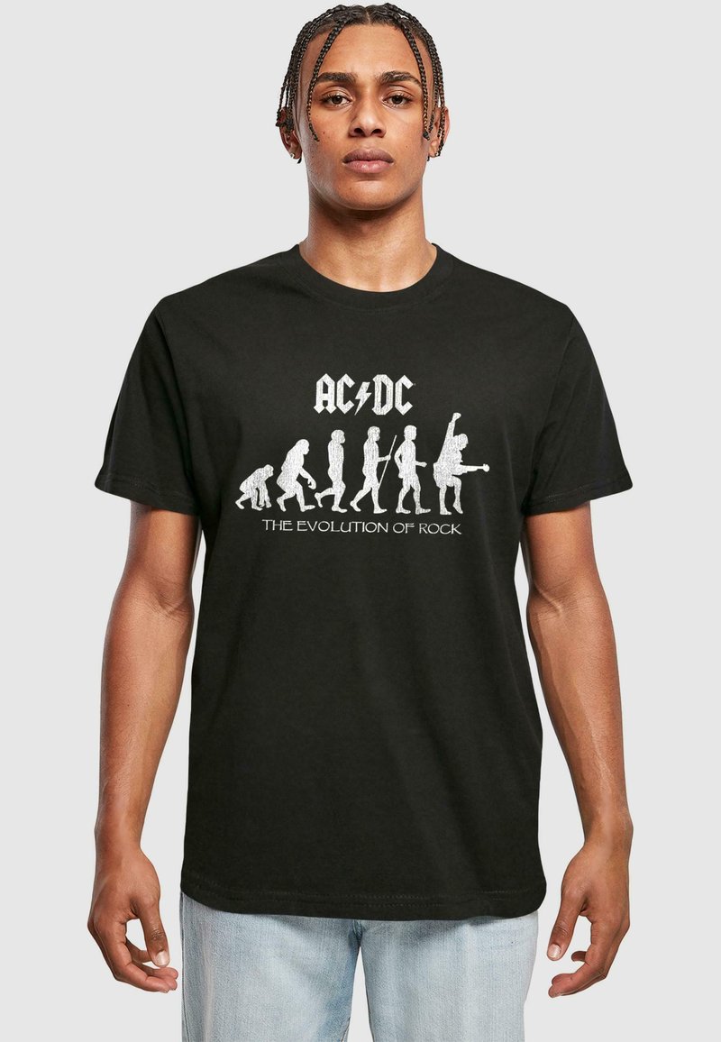 Svart t-skjorte med teksten "AC/DC" og "The Evolution of Rock" i hvitt, med grafiske silhuetter som viser menneskelig evolusjon til rockemusikere.