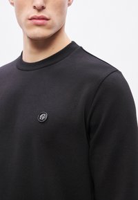 Čierna mikina s okrúhlym výstrihom a rebrovanými manžetami. Na hrudi má malý čierny logotyp, vyrobená je z hladkej, mäkkej látky.