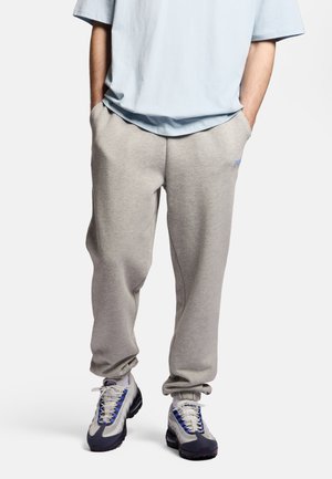 Osoba nosząca jasnoszare spodnie dresowe z elastycznymi mankietami, jasnoniebieską oversize koszulkę i białe sneakersy z niebieskimi i czarnymi akcentami.