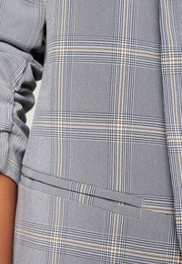 Blazer à carreaux gris avec fines rayures verticales, présentant une poche verticale et des accents dorés contrastants dans le motif. Tissu texturé.