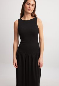 Must esine rihmata, millel on väljakujunenud rinnaosa ja voolav, kokku pandud seelik. Materjal näib olevat tekstureeritud ja pehme, ümara kaelusega.