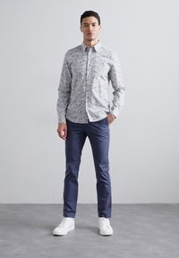 Camicia bianca a maniche lunghe con un motivo floreale, abbinata a pantaloni blu scuro e sneakers bianche. La camicia presenta un colletto classico.