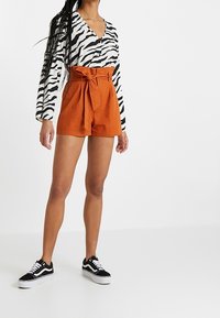 Blusa a maniche lunghe con stampa zebra e scollo a V, abbinata a pantaloni shorts arancioni a vita alta con dettaglio a fiocco, indossata con sneaker nere.