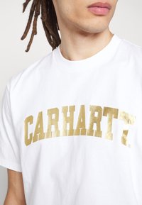 Valkoinen puuvillapaita, jossa on suuri, metallinhohtoinen kultainen "CARHARTT" -logo. Pyöreä kaula-aukko ja lyhyet hihat, sileä pinta.