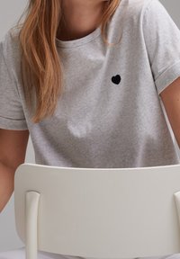 Šedé bavlněné tričko s jemnou texturou, zdobené malou černou výšivkou srdce na levém prsu, krátké rukávy a kulatý výstřih.