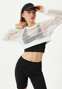 Czarny stanik sportowy pod białym, siateczkowym, krótkim swetrem i czarnymi szortami rowerowymi o wysokim stanie. Modelka w czarnej czapce przeciwsłonecznej. Gładkie tekstury.