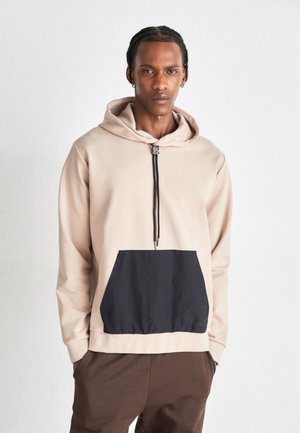 Haut à capuche beige avec une poche avant noire, un cordon de serrage au niveau de la capuche et un col zippé. Texture douce, design décontracté et coupe relax. 