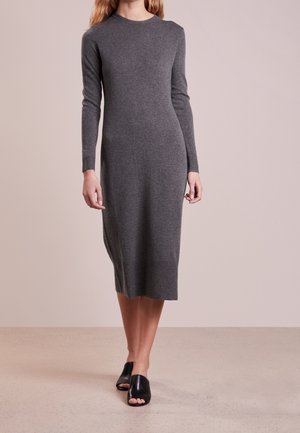 Kobieta w szarej dzianinowej sukience z długim rękawem i sięgającej kolan oraz czarnych otwartych klapkach, stoi na jednolitym beżowym tle.