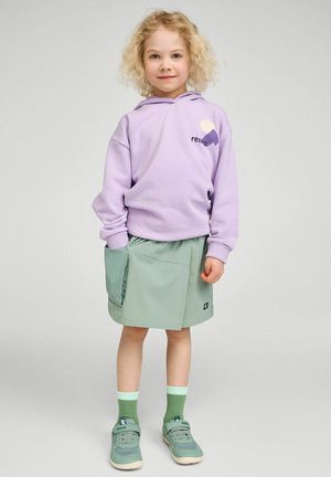 Małe dziecko z kręconymi blond włosami, ubrane w lawendową bluzę z kapturem, jasnooliwkową spódniczkę z dużą kieszenią, zielone skarpetki i zielone trampki, stojące.