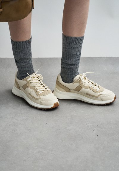 Beige Sneaker mit Wildlederakzenten, weißen Sohlen und cremefarbenen Schnürsenkeln. Kombiniert mit grauen gerippten Socken, die ein strukturiertes Muster aufweisen.