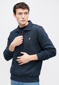 Tumesinine pullover-kampsun poollukuga kraega, soonikkoes varrukaotsad ja alumine ääreosa. Vasakul rinnal on väike kollane linnu logo.