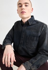Camicia button-up in denim scuro con taschino sul petto, texture vissuta e colletto classico. Il modello posa con le mani appoggiate sulle ginocchia.