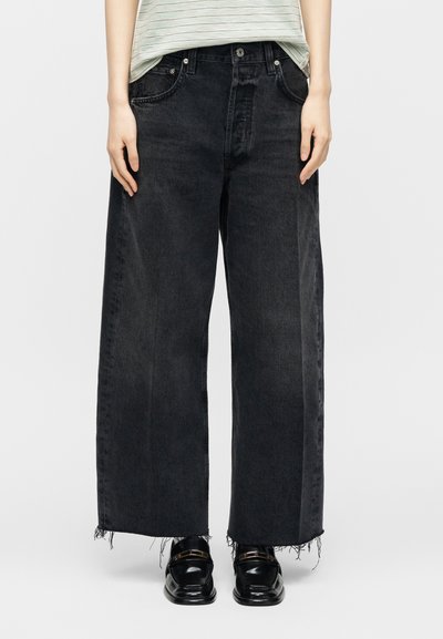 Persoon die losse zwarte denim jeans draagt met rafelige zomen, een ingestopt gestreept overhemd en zwarte glanzende loafers met gouden accenten.