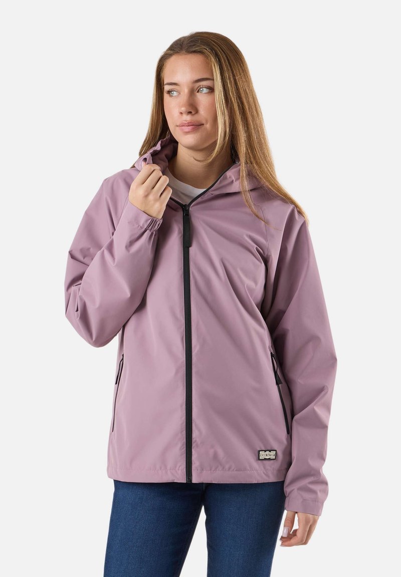 Młoda kobieta z długimi włosami w mauve kurtce zapinanej na zamek z kieszeniami, trzymająca kołnierz, patrząca na bok na jednolitym tle.