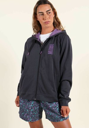 Femme portant un sweat à capuche noir zippé avec des accents violets et un short à motifs, debout devant un fond clair, regardant sur le côté.