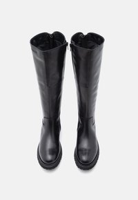 Pár čiernych kožených vysokých čižiem s bočnými zipsami a robustnými gumovými podrážkami, vystavených vo vzpriamenej polohe na bielom pozadí.