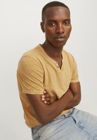 Camisa de manga corta, estilo henley, en un color mostaza claro, con un tejido texturizado y dos botones en el cuello. Llevada con jeans de mezclilla.