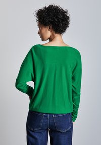 Grüner Pullover mit langen Ärmeln, im lässigen Schnitt und weitem Ausschnitt. Die Rückseite ist glatt und unauffällig, kombiniert mit dunkelblauen Jeans.