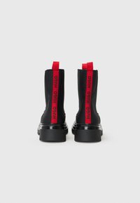 Botas de tornozelo pretas com acabamento mate e solas de plataforma grossas. Apresentam alças de tecido vermelho com a marca "HUGO" de cada lado.