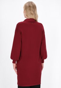 Abito lavorato a maglia bordeaux con vestibilità comoda, dotato di un colletto alto, maniche a tre quarti e polsini a coste, che arriva a metà coscia.