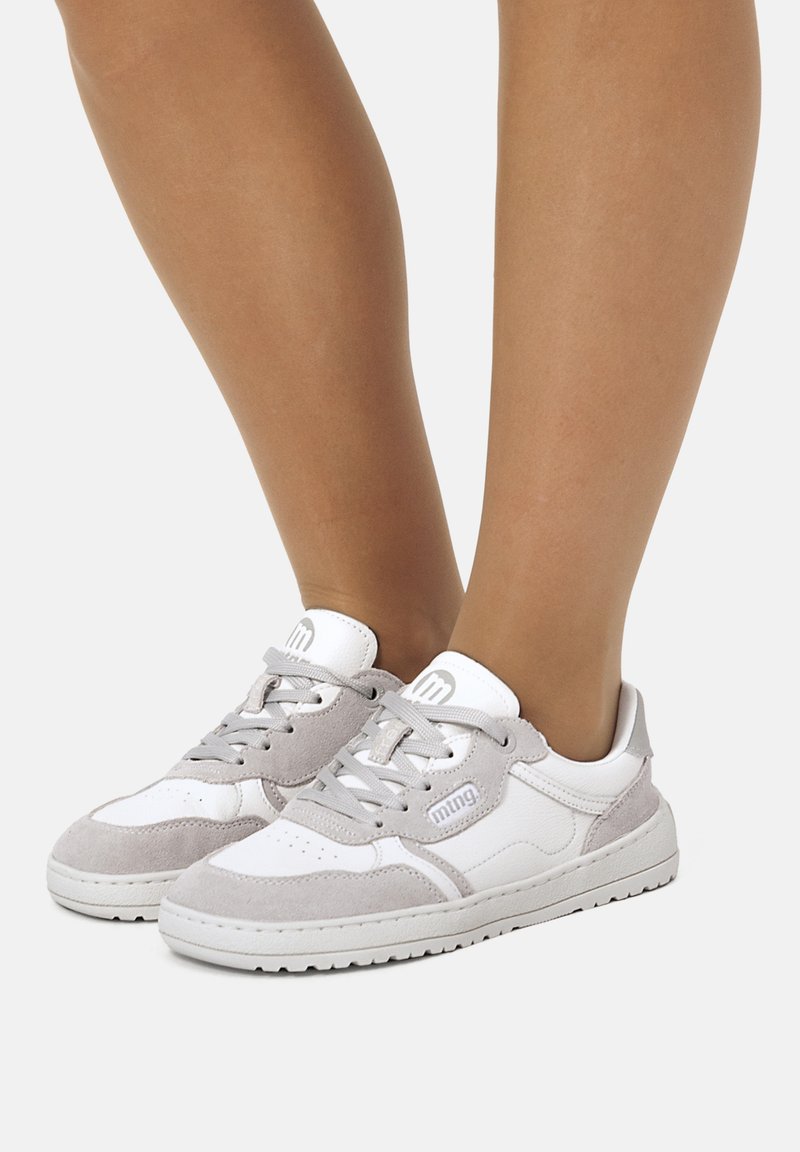 Persona vistiendo zapatillas bajas de color blanco y gris claro con cordones, de pie sobre un fondo blanco.