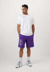 Hvid t-shirt med kortærmer og cirkulært logo, parret med lilla shorts med et Lakers-grafik og logo, samt grå sneakers.