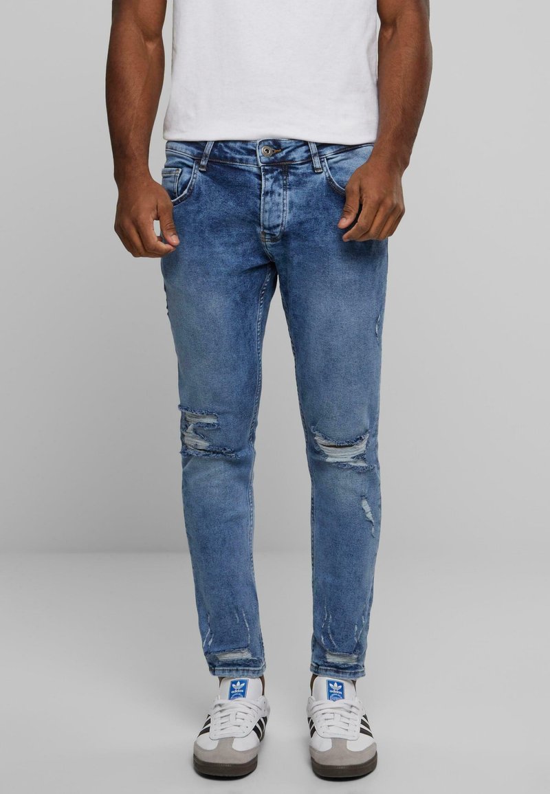 Világoskék, slim fit farmernadrág. Térdén és zsebein kopott hatású. Fehér tornacipővel viselve.