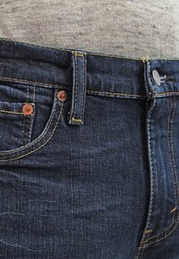 Tummansiniset farkut, joissa on kupariset niitit, keltaiset ompeleet ja perinteinen viiden taskun malli. Sileä pinta ja istuva muoto.