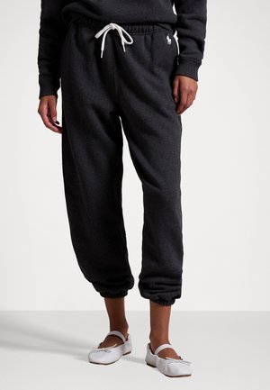 Personne portant un pantalon de survêtement gris foncé avec des poignets élastiques et un cordon blanc, associé à des ballerines blanches sur un fond uni.