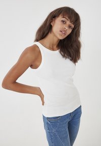 Débardeur blanc à côtes avec un col rond et des emmanchures larges, associé à un jean bleu. Le tissu est doux et ajusté.